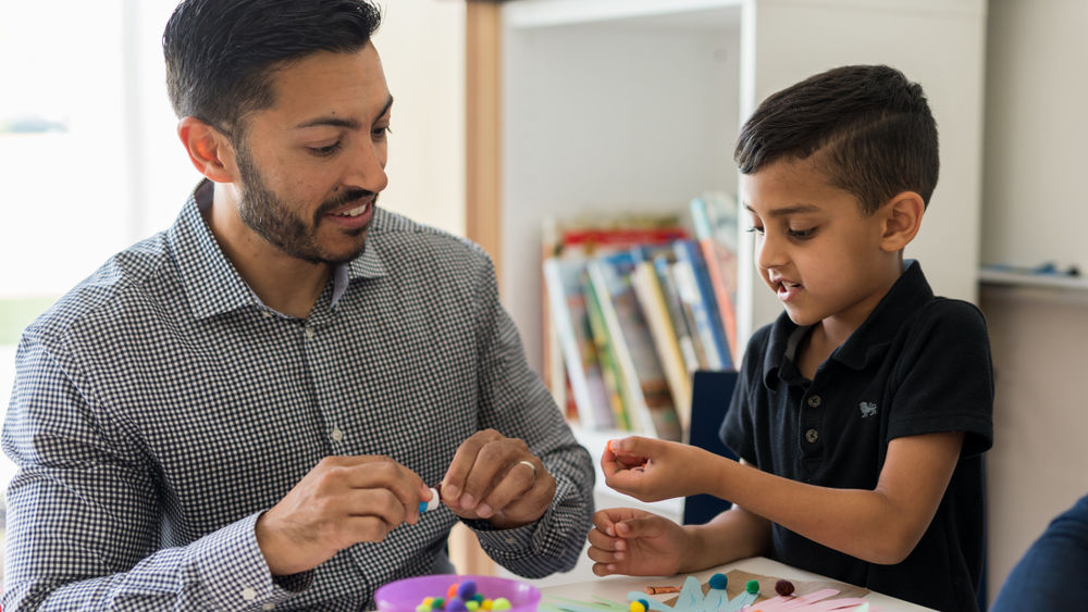 A man and a little boy, both holding art materials, look at each other’s hands.