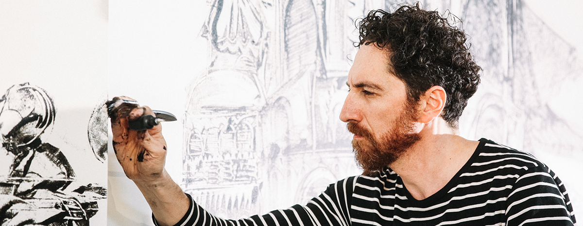 Artist Kevork Mourad, in a black-and-white- striped T shirt, touches a paintbrush to a wall decorated with his black-and-white artwork