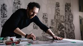 A man in a black long sleeve shirt is leaning over a table drawing with charcoal.