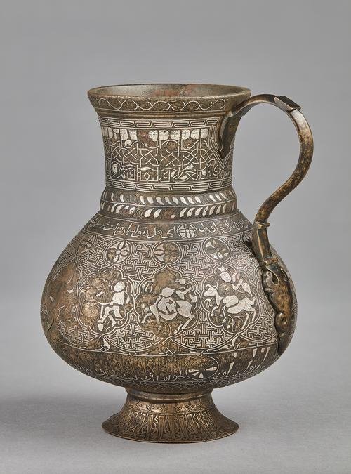 A pear-shaped brass jug with handle on a gently splayed foot, with silver inlaid decoration comprising of interlinked medallions containing representations of the zodiac signs, on a T-fret pattern background, between two inscriptive bands; the neck with a frieze of human-headed Kufic inscription. 